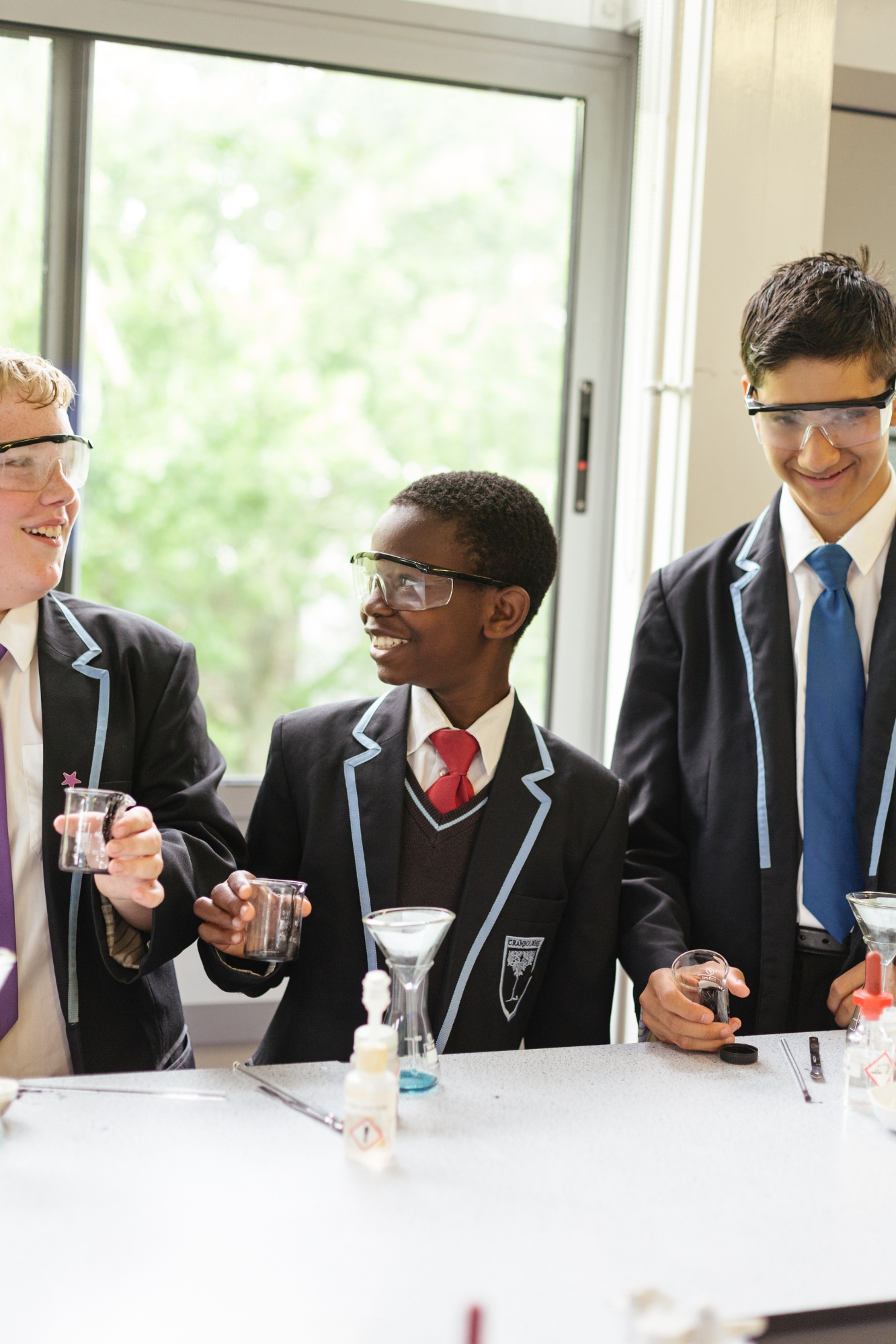 3 male students, smiling and wearing safety googles. Hholding science equipment